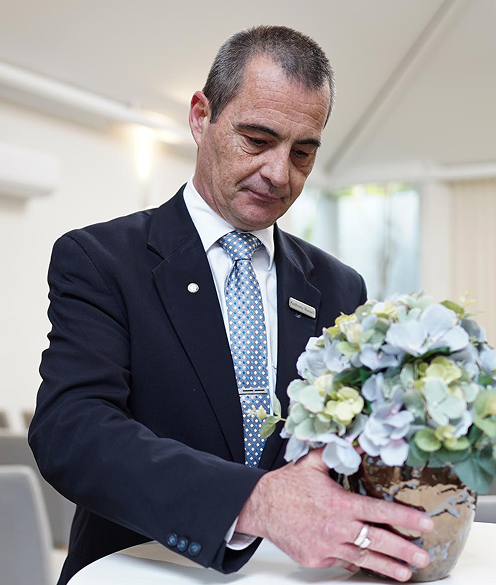 Image of a man dressed in a funneral arranger's uniform is placing a vase of flowers on a tall table