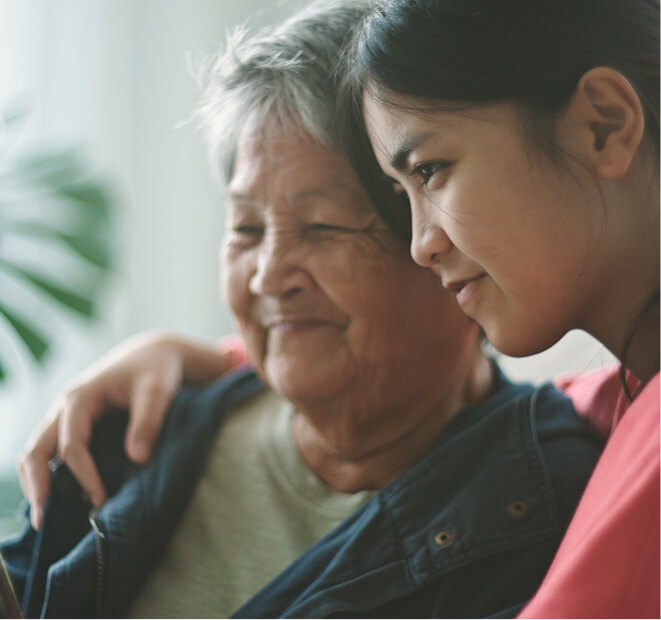 An image of a two women in a side hug. One woman looks like an eldery woman and a younger woman is giving her a side hug and they are both smiling